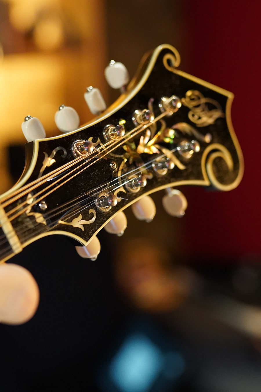 A hand on the neck of an F-style mandolin, its headstock in sharp focus: ivory tuner buttons, abalone inlay, and the scroll.